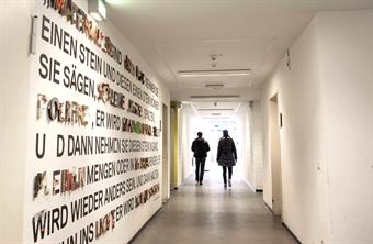 Zwei Studenten gehen einen hellen Flur der HSD hinunter. Die linke Wand ist mit Schriftzügen aus verschiedenen Materialien geschmückt.
